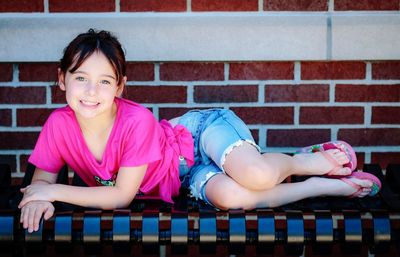 Portrait of smiling girl lying on bench against brick wall