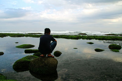 Rear view of boy crouching on rock in sea against sky