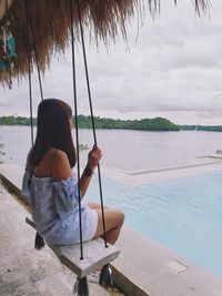 Woman sitting swing while looking at lake against cloudy sky