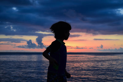 Boy standing at beach against cloudy sky during sunset