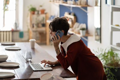 Young female pottery business owner using laptop talking with client by phone, checking online order