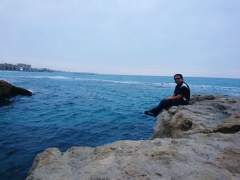 Man sitting on rock by sea against clear sky