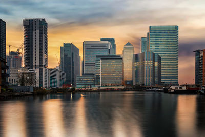 Modern buildings by river against sky during sunset