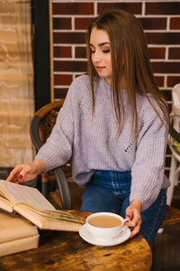 A beautiful girl in a gray knitted sweater drinks coffee and reads a book in a cozy cafe.
