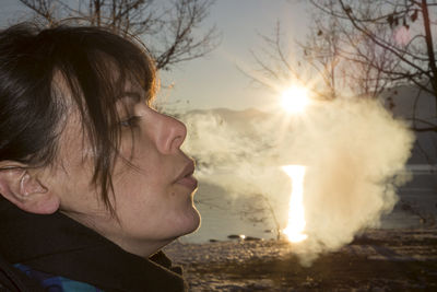 Side view of woman exhaling smoke while standing against lake