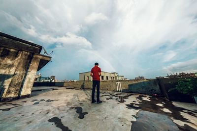 Rear view of man standing at building terrace against cloudy sky