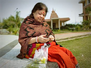 Senior woman holding leaves while sitting on footpath against temple
