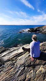 Rear view of woman sitting on rock looking at sea shore against sky