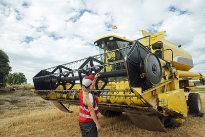 Back view of anonymous female worker in uniform examining combine harvester while working in agricultural field in countryside on summer day