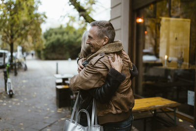 Happy man embracing woman while standing sidewalk near restaurant