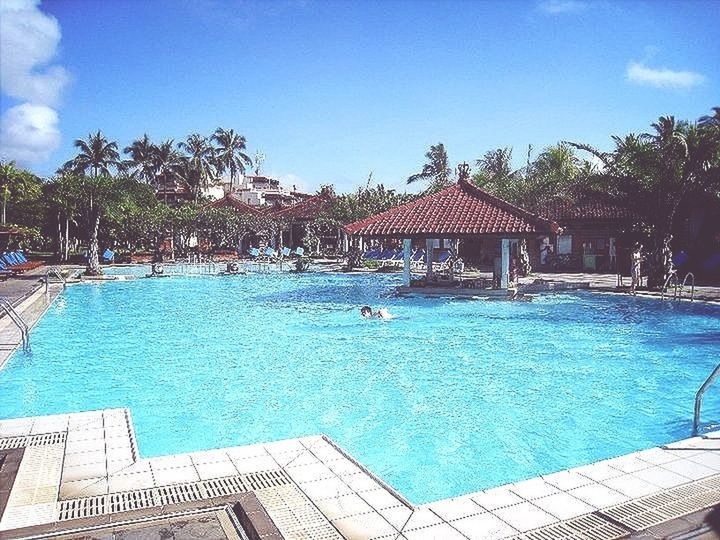 water, swimming pool, tree, building exterior, blue, built structure, architecture, turquoise colored, sky, sunlight, tourist resort, day, sea, palm tree, vacations, house, incidental people, outdoors, nature