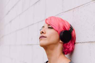 Young female with bright pink hair listening to music with headphones while standing near white wall with eyes closed