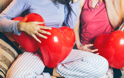 Midsection of female friends with heart shape balloons sitting on sofa at home