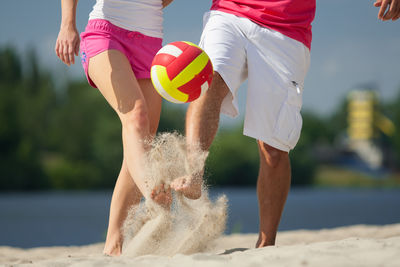 Low section of couple kicking volley ball while playing on sand at beach