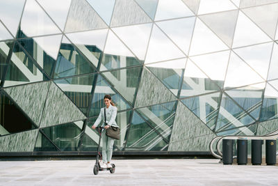 Happy female entrepreneur in smart casual style clothing riding electric scooter near glass building in downtown while commuting to work and looking down