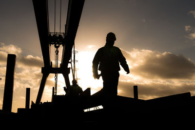 Silhouette worker by metallic structure against cloudy sky during sunset