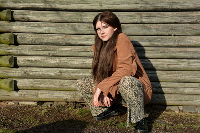 Young girl crouches on the floor in front of old wooden planks in the sun and looks forward