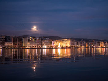 Illuminated buildings by river against sky at night