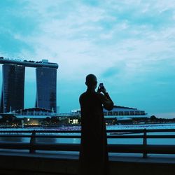 Rear view of man photographing on mobile phone against sky in city