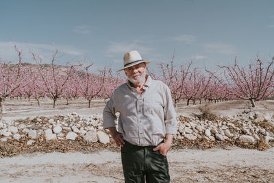 Content mature male farmer in hat looking at camera while standing near blossoming apricot trees in orchard