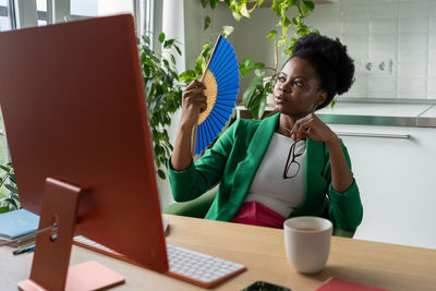 African businesswoman uses hand fan sits at desk with computer in summer day, suffers from heat.