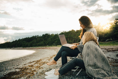Woman wrapped in blanket looking at friend using laptop on sea shore