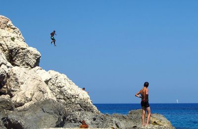 Men standing on cliff in front of sea against clear blue sky