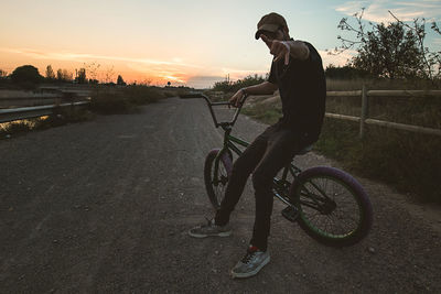 Man gesturing shaka sign while sitting on bicycle against sky during sunset