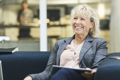 Happy businesswoman looking away while holding digital tablet on sofa at office lobby