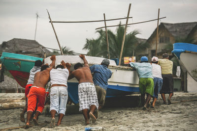 Rear view of people pushing boat at sandy beach