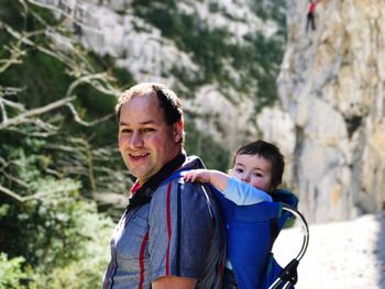 Side view portrait of happy father carrying son in baby carrier against mountains