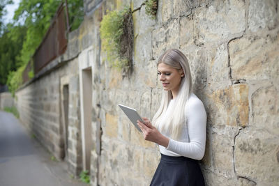 Side view of young woman using phone while standing against wall