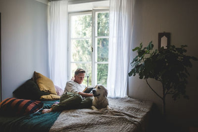 Full length of retired man reading book while resting with dog on bed at home