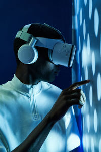 Side view of concentrated young african american in white t shirt and modern vr goggles pointing and touching screen while interacting with virtual object in dark studio with neon illumination