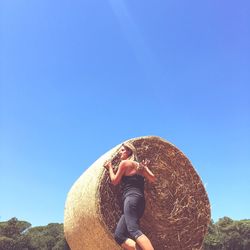 Low angle view of woman leaning on hay bale against clear blue sky during sunny day