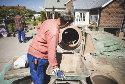 Side view of senior woman using cement mixer while man standing in background at yard