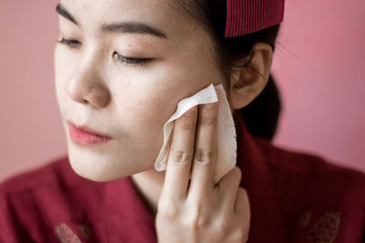 Close-up of woman cleaning face by tissue against coral background