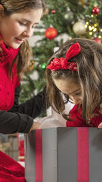 Two girls in different age open a christmas present, blurred christmas  tree as background.