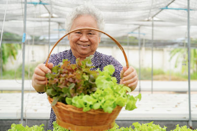 Portrait of smiling senior woman holding basket with leaf vegetables in greenhouse