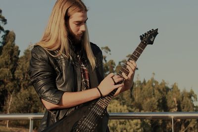 Young man holding guitar against sky