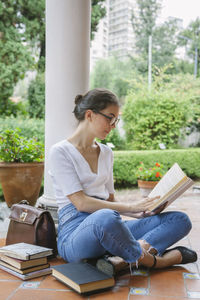 Young woman reading book while sitting on floor against plants