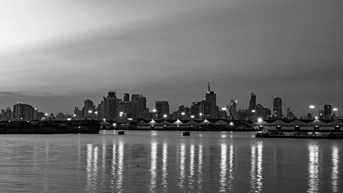 Illuminated buildings by river against sky in city