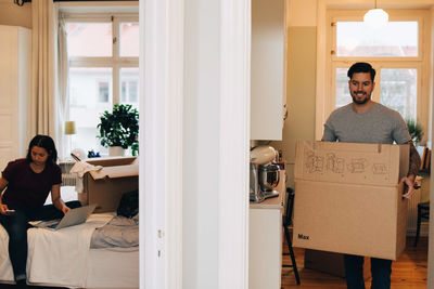 Man carrying box at doorway while woman using laptop in bedroom