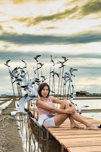 Portrait of mid adult woman sitting by sculptures on pier over lake against cloudy sky during sunset