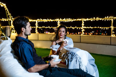 Young couple having coffee while relaxing on illuminated terrace at night