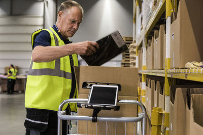 Side view of senior male worker packing merchandise in cardboard box on cart at distribution warehouse