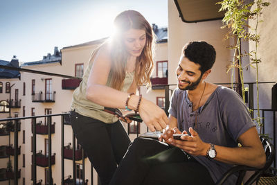 Happy couple using smart phone while reading guidebook on balcony