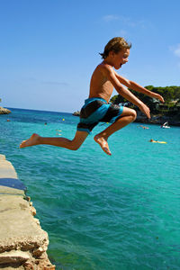 Full length of shirtless boy jumping in sea against blue sky