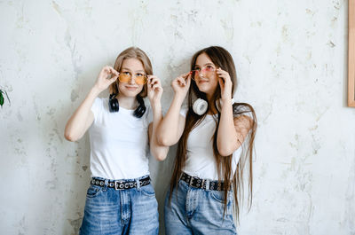 Two teenage girls stand near a white wall in funny sunglasses. . headphones hang on their necks