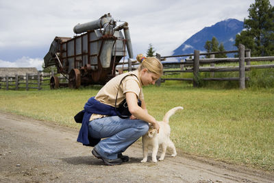 Side view of young woman with cat crouching on field against cloudy sky